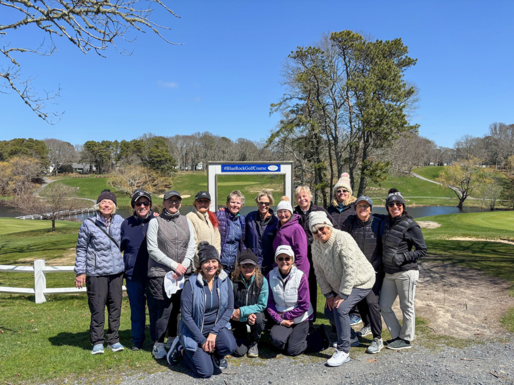 A group of 15 people in jackets and hats pose together in front of a golf course sign under a bright blue sky, with trees and green grass in the background.