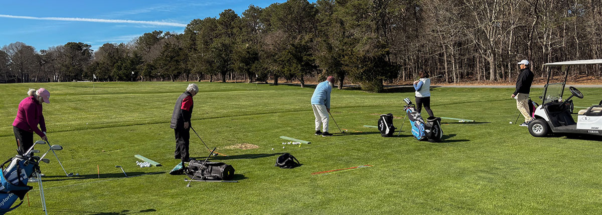 Five people practice golf on a grassy driving range under a clear blue sky, with golf bags and clubs nearby. A golf cart is parked on the right, and trees line the background.