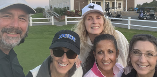 Five adults, four women and one man, smile for a selfie on a golf course. Two women wear caps, and golf carts and a building are visible in the background. The group appears happy and relaxed outdoors.