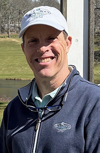 A man wearing a white baseball cap, sunglasses around his neck, and a navy blue zip-up jacket stands outdoors, smiling, with a grassy field and trees in the background.