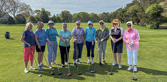 Nine women stand in a row on a golf course, each holding a golf club, smiling at the camera. The background features green grass, trees, and a partly cloudy sky.