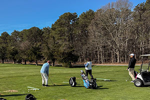 Three people stand on a golf course, two preparing to hit balls while one observes. Golf bags and a cart are nearby, with trees and a blue sky in the background.