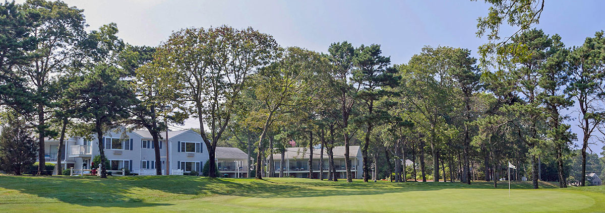 View of the first hole at Blue Rock Golf Course, with neatly trimmed grass, surrounded by tall trees, and white buildings in the background under a partly cloudy sky.