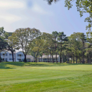 View of the first hole at Blue Rock Golf Course, with neatly trimmed grass, surrounded by tall trees, and white buildings in the background under a partly cloudy sky.