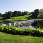 A white bridge crosses a calm pond at Blue Rock Golf Course, surrounded by green grass and trees under a clear blue sky, with rolling hills and sand bunkers in the background.