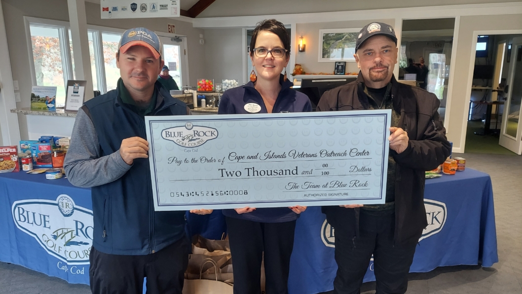 Three people stand indoors holding a large ceremonial check for two thousand dollars made out to the Cape and Islands Veterans Outreach Center from the Town of Blue Rock. Tables with canned goods and Blue Rock signs are visible behind them.