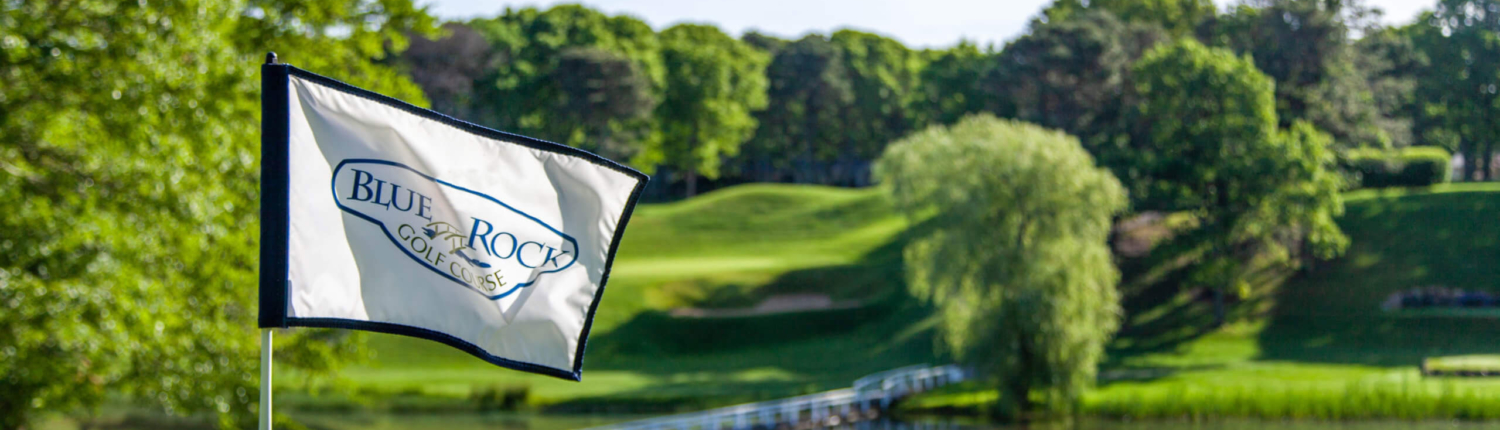 A Blue Rock Golf Course flag waves on a lush green, overlooking a tree-lined fairway with a small pond, footbridge, and the bright blue sky—all capturing the charm of Blue Rock Golf Course.