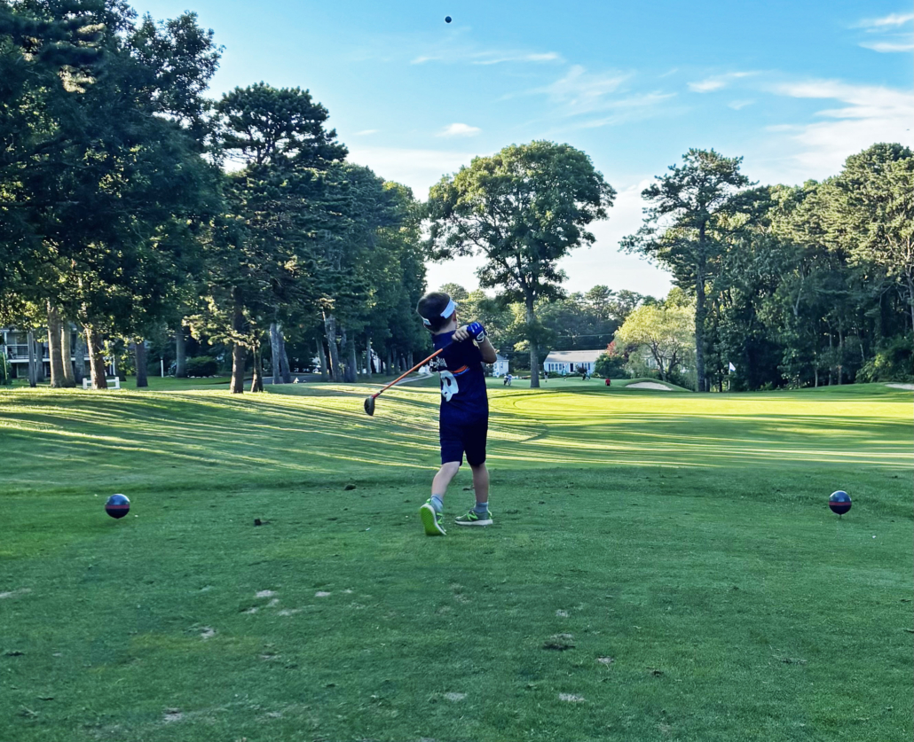 A young boy wearing a blue shirt and shorts swings a golf club on a green golf course, surrounded by trees under a blue sky.
