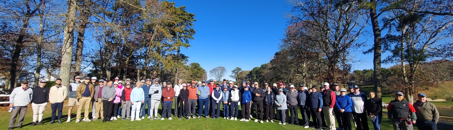 A large group of people wearing golf attire stand together on a golf course with trees and clear blue sky in the background, posing for a group photo.