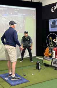 Two men are in an indoor golf simulator. One stands ready to swing a club while the other, facing him, appears to be coaching. Golf equipment and monitors are visible, with a virtual golf course projected in the background.