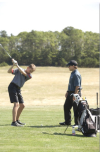 Two young golfers stand on a grassy golf course; one is swinging a club while the other watches. A golf bag and equipment are nearby, with trees and open field in the background.
