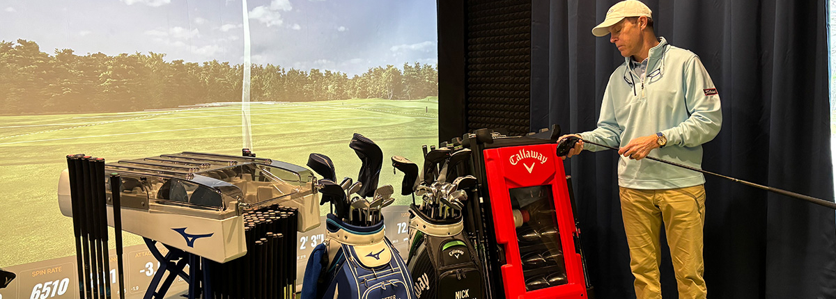 A man in golf attire stands indoors beside several golf bags and a club-fitting machine, holding a golf club. A large screen behind him displays a virtual golf course.