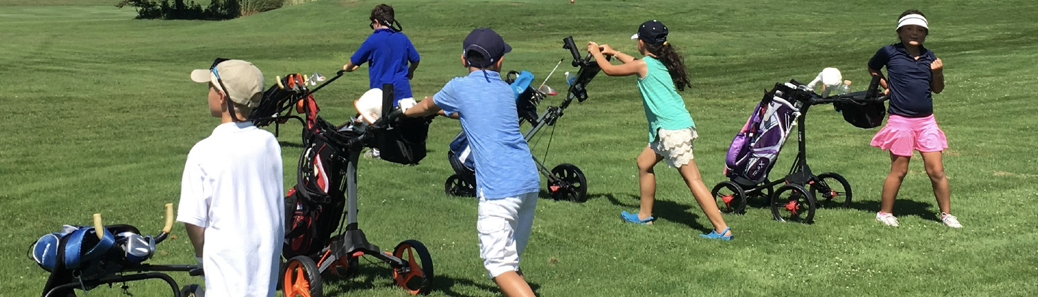 A group of children on a grassy golf course push golf trolleys and carry bags under a bright, sunny sky. Some wear hats and colorful outfits as they walk and prepare to play golf.