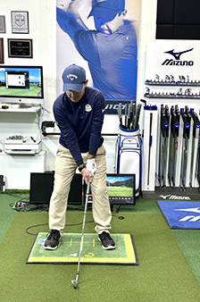 A golfer wearing a blue cap and shirt with beige pants stands ready to swing in an indoor golf fitting room, surrounded by golf clubs, equipment, and golf brand posters.