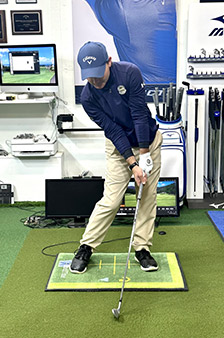 A golfer wearing a blue cap and shirt, tan pants, and black shoes prepares to hit a golf ball indoors on a practice mat, surrounded by golf equipment, monitors, and a golf-themed wall display.