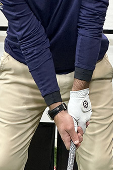 A person wearing a navy long-sleeve shirt and beige pants holds a golf club, showing a close-up of their grip. They are wearing a white golf glove and a black wristwatch.