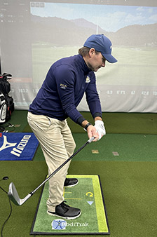 A golfer wearing a blue cap and jacket prepares to swing indoors, standing on a golf mat with a club in hand. A large screen behind displays a virtual golf course.