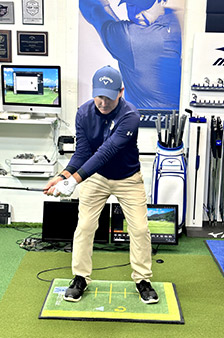 A man in a blue cap and shirt stands in a golf stance on an indoor golfing mat, holding a club. Golf equipment, awards, and a large photo of a golfer are visible in the background.