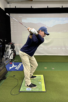 A person in a navy shirt, light pants, and a cap practices a golf swing indoors, standing on a green mat with a golf simulator screen displaying a course in the background.