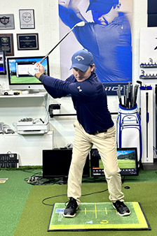 A person in golf attire practices their swing indoors on a golf mat, with golf equipment, awards, and screens visible in the background. A large poster of a golfer in mid-swing hangs on the wall behind them.