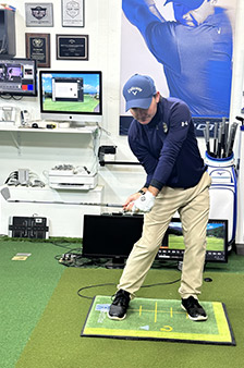 A man in golf attire practices his swing indoors on a green mat, surrounded by golf equipment, monitors, and award plaques on the wall.