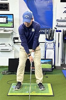 A golfer wearing a blue cap and shirt stands indoors on a golf mat, holding a club and preparing to swing. Golf equipment, monitors, and screens are visible in the background.