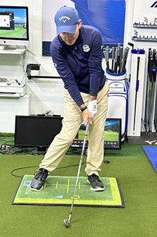 A golfer in a navy cap and jacket prepares to swing an iron club indoors, standing on a green mat with yellow lines. Golf clubs and training equipment are visible in the background.