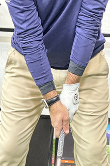 A person wearing a blue long-sleeve shirt and khaki pants holds a golf club with both hands, wearing a white glove on the left hand, standing in a ready stance.