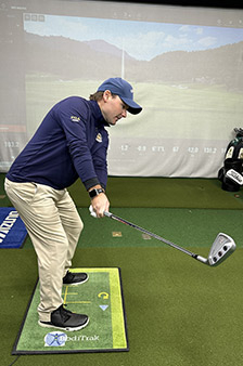 A person in a blue long-sleeve shirt, khaki pants, and a cap is practicing a golf swing indoors on a green golfing mat, with a golf simulator screen displaying a virtual golf course in the background.
