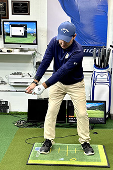 A man wearing a blue cap and golf attire is practicing his golf swing indoors on a mat, with golf equipment, computers, and monitors in the background displaying a golf simulation.