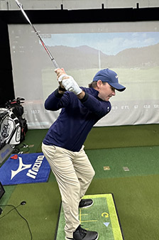 A person wearing a blue cap and navy top practices a golf swing indoors on an artificial turf mat, with a golf simulator screen displaying a golf course in the background.
