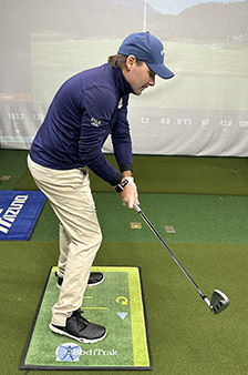 A man in golf attire stands indoors on an artificial turf mat, holding a golf club and preparing to take a swing. A golf course scene is projected on the wall behind him.