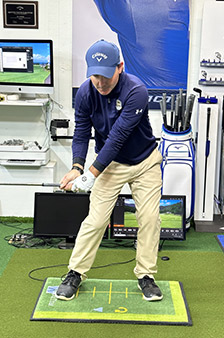A man wearing a blue cap, navy long-sleeve shirt, and khaki pants practices a golf swing indoors on a green mat, surrounded by golf equipment, monitors, and training aids.