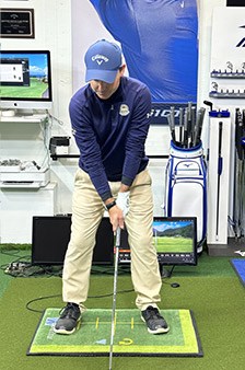 A golfer wearing a blue hat and shirt stands indoors on a green mat, preparing to swing a club, with golf equipment, monitors, and a golf bag in the background.
