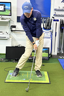 A man in golf attire prepares to swing a golf club indoors, standing on a green mat surrounded by golf equipment, computer monitors, and clubs in a training facility.