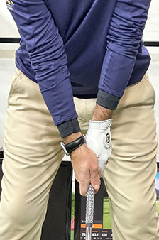 A person wearing a blue long-sleeve shirt and beige pants is gripping a golf club, preparing for a swing, with both hands on the club and a golf glove on the left hand.