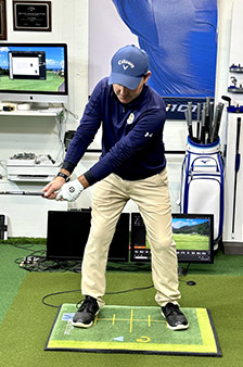 A man in golf attire stands indoors on a practice mat, holding a golf club mid-swing. Behind him are screens, golf equipment, and sports memorabilia. He appears to be practicing his golf technique.