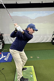 A man in a blue long-sleeve shirt, beige pants, and a cap is mid-backswing with a golf club on an indoor golf simulator, standing on a mat with a projected golf course in the background.