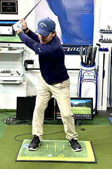 A golfer in a blue shirt and khaki pants prepares to swing indoors, standing on a practice mat with alignment lines. Golf equipment, monitors, and golf bags are visible in the background.