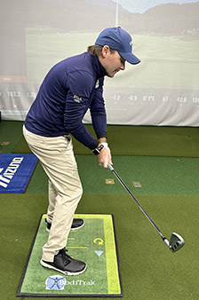 A man dressed in a blue jacket, beige pants, and a blue cap prepares to swing a golf club indoors, standing on a yellow and green mat labeled BodiTrak. A golf simulator screen displays a course in the background.