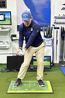 A golfer in a blue cap and navy shirt stands indoors on a green mat, preparing to swing a club. Golf equipment, monitors, and screens are visible in the background, suggesting a training or simulation area.