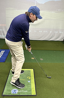 A man wearing a blue cap, navy jacket, and beige pants stands on a golf training mat indoors, preparing to swing a golf club on an artificial turf surface.