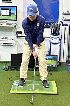 A golfer stands indoors on a practice mat, holding a club and preparing to swing. He wears a blue cap and shirt, beige pants, and black shoes. Golf equipment, monitors, and bags are visible in the background.