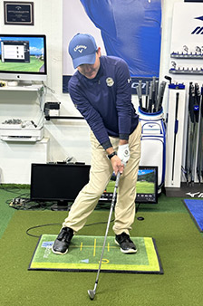 A person wearing a blue hat and shirt, tan pants, and black shoes demonstrates a golf swing indoors on an artificial turf mat, holding a golf club and looking down at the ball. Golf equipment and monitors are visible in the background.