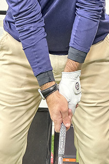 A person wearing a blue long-sleeve shirt, khaki pants, and a white golf glove grips a golf club, preparing for a swing. The focus is on their hands and the upper part of the club.