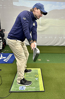 A man in a blue jacket and cap practices his golf swing indoors, standing on a golf mat with alignment markings. He holds a golf club and appears focused, with golf equipment and a screen in the background.