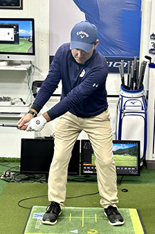 A golfer in a blue shirt, khaki pants, and blue cap practices a golf swing indoors on a mat, surrounded by monitors, golf clubs, and training equipment.