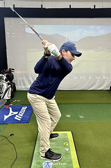 A man in a navy shirt, khaki pants, and a blue cap is holding a golf club in mid-swing at an indoor golf simulation facility, with a projected golf course in the background.