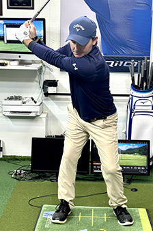 A man in a navy shirt, khaki pants, and a blue cap is holding a golf club at the top of his backswing indoors, standing on a mat with golf training equipment and monitors around him.