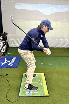 A golfer wearing a blue jacket and cap practices his swing indoors on a green mat, holding a golf club mid-swing. A golf bag and virtual golf simulator screen are visible in the background.